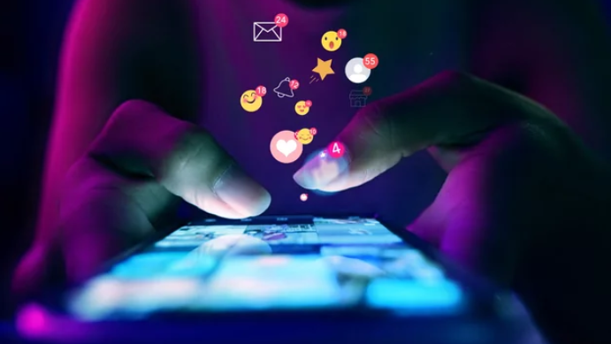 Une personne tient un téléphone dans la pénombre ; l’écran lumineux se détache nettement dans l’obscurité. Des émojis — cœurs, icônes de réseaux sociaux, réactions — semblent jaillir de l’appareil, symbolisant les interactions et la dynamique des réseaux sociaux.